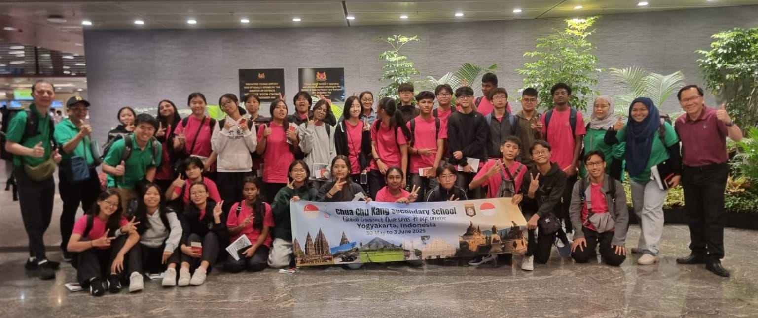 A large group of students in pink shirts pose at an airport holding a Chua Chu Kang Secondary School banner for a trip to Yogyakarta, Indonesia.