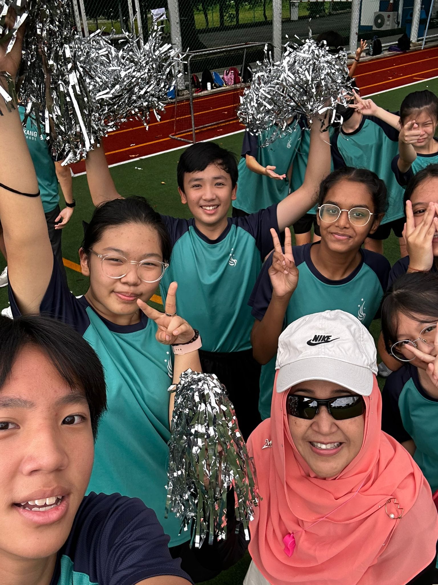 Group of students in teal uniforms holding silver pom-poms, posing with peace signs on a sports field.