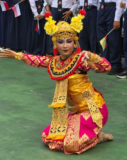 A performer in gold and pink traditional Balinese costume and headdress dances on a green floor before students.