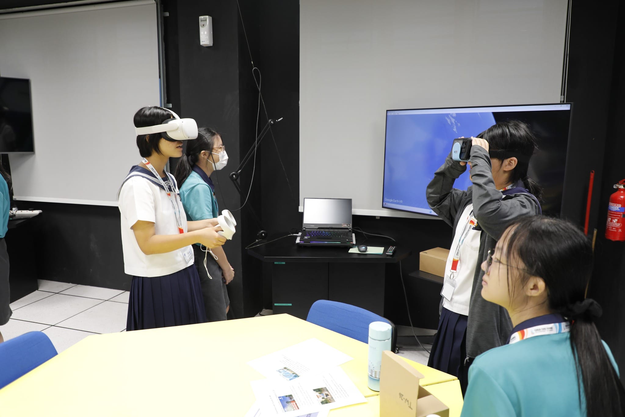 Students in school uniforms use VR headsets in a classroom with a large screen showing Google Earth VR.
