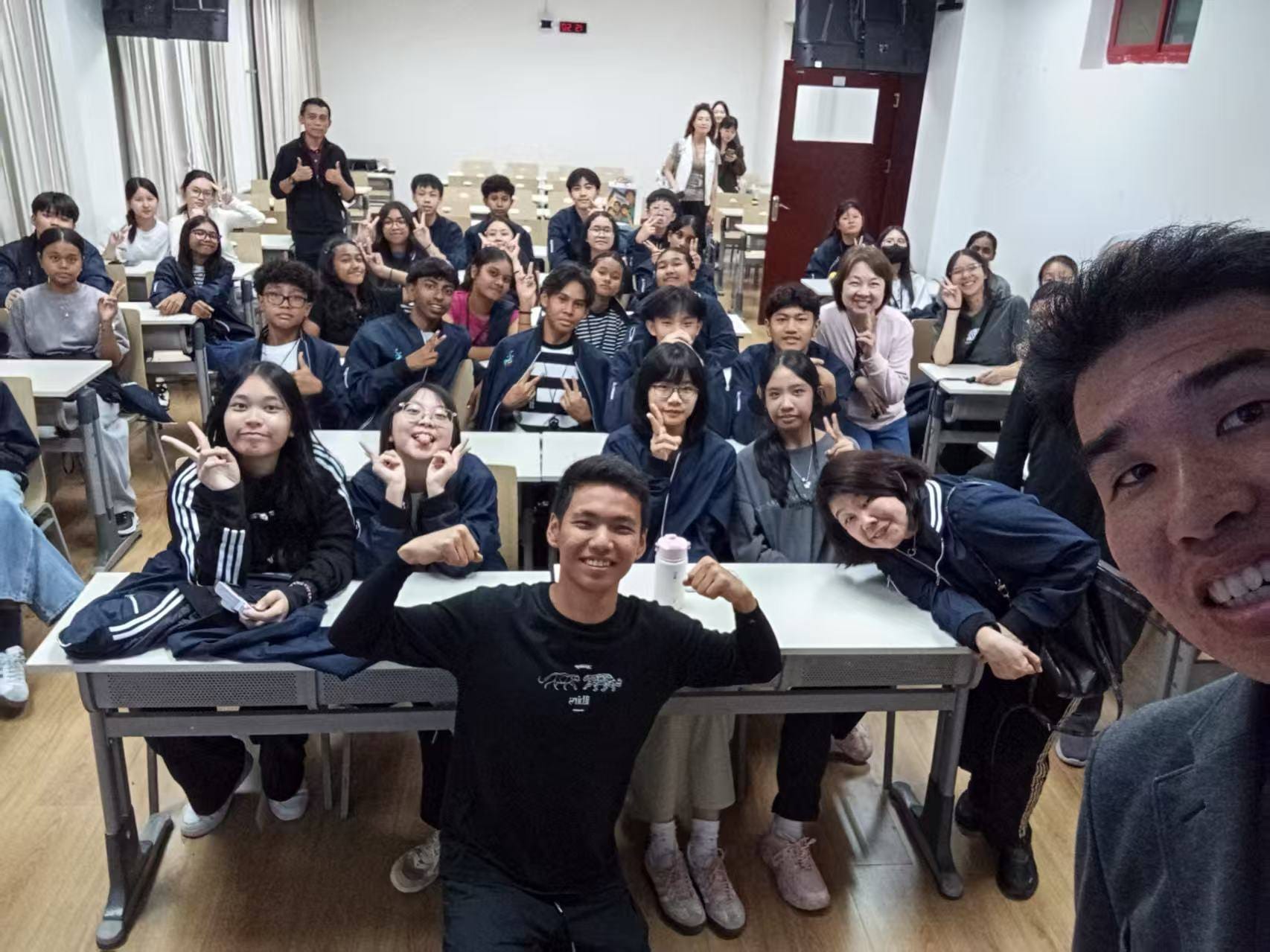 A large group of students and teachers pose smiling in a classroom, many making peace signs toward the camera.