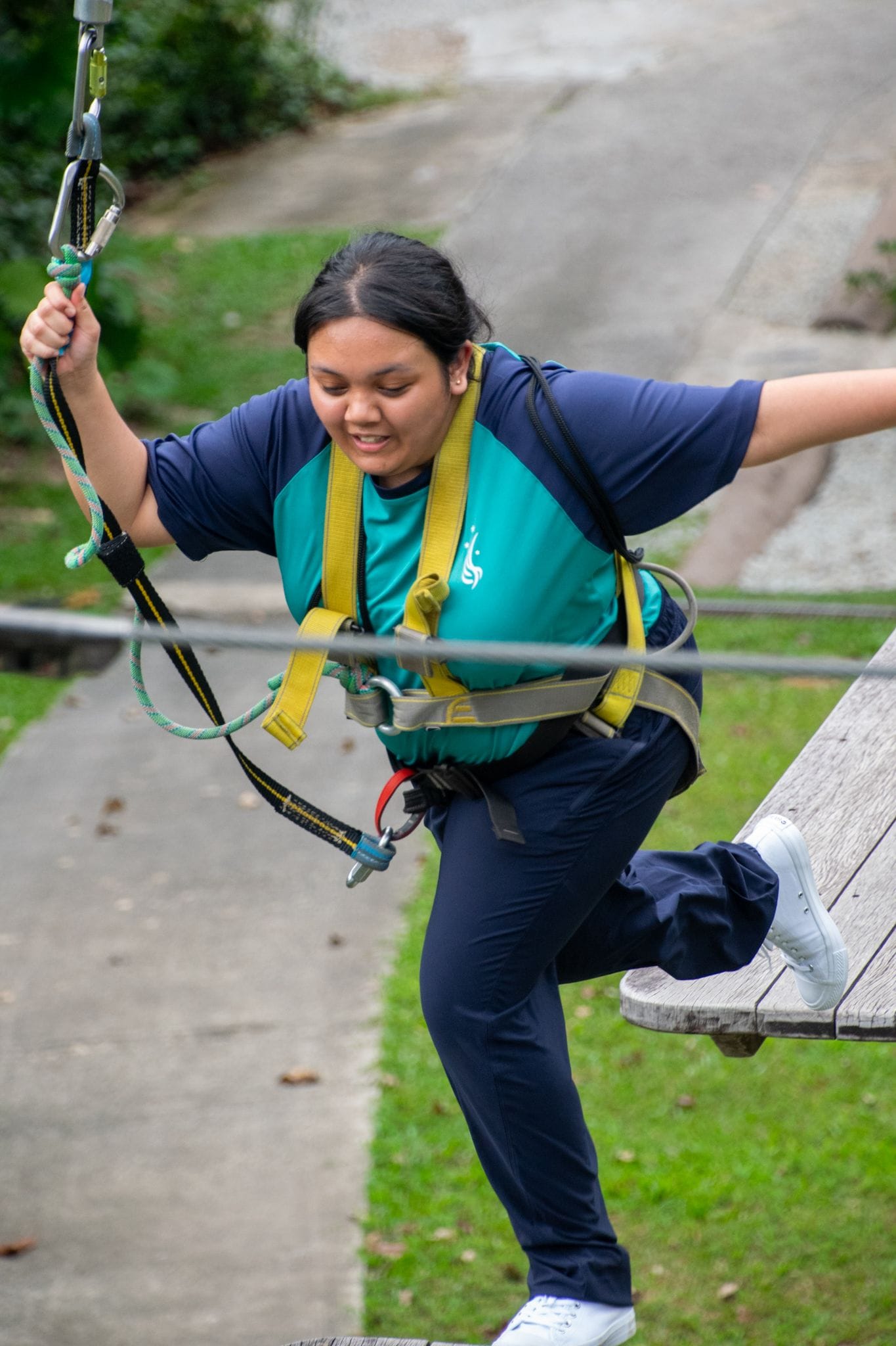 A young woman wearing a yellow safety harness grips a rope while stepping off a wooden platform outdoors.