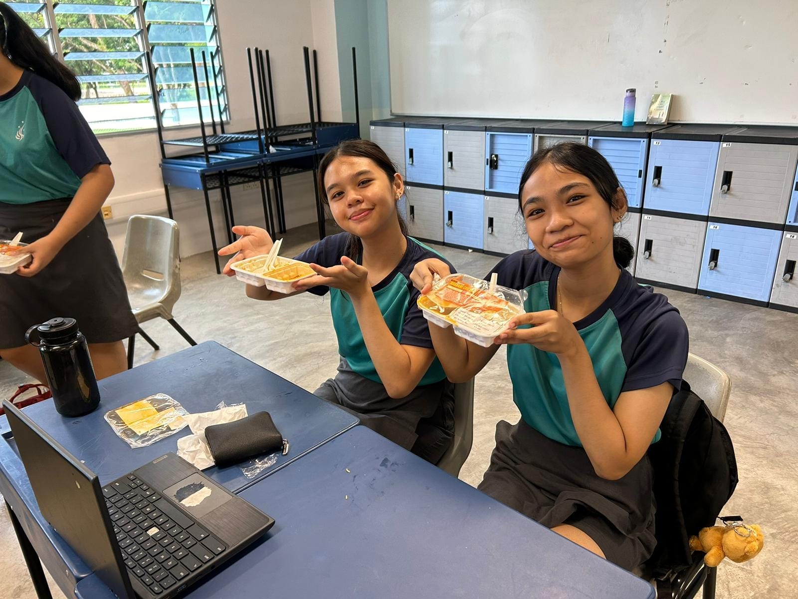 Two smiling female students in matching uniforms hold up plastic food containers at a desk in a classroom.