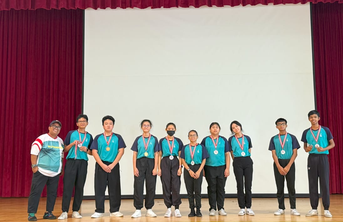 Ten people stand on a stage wearing medals, most in matching teal uniforms, in front of red curtains and a white screen.