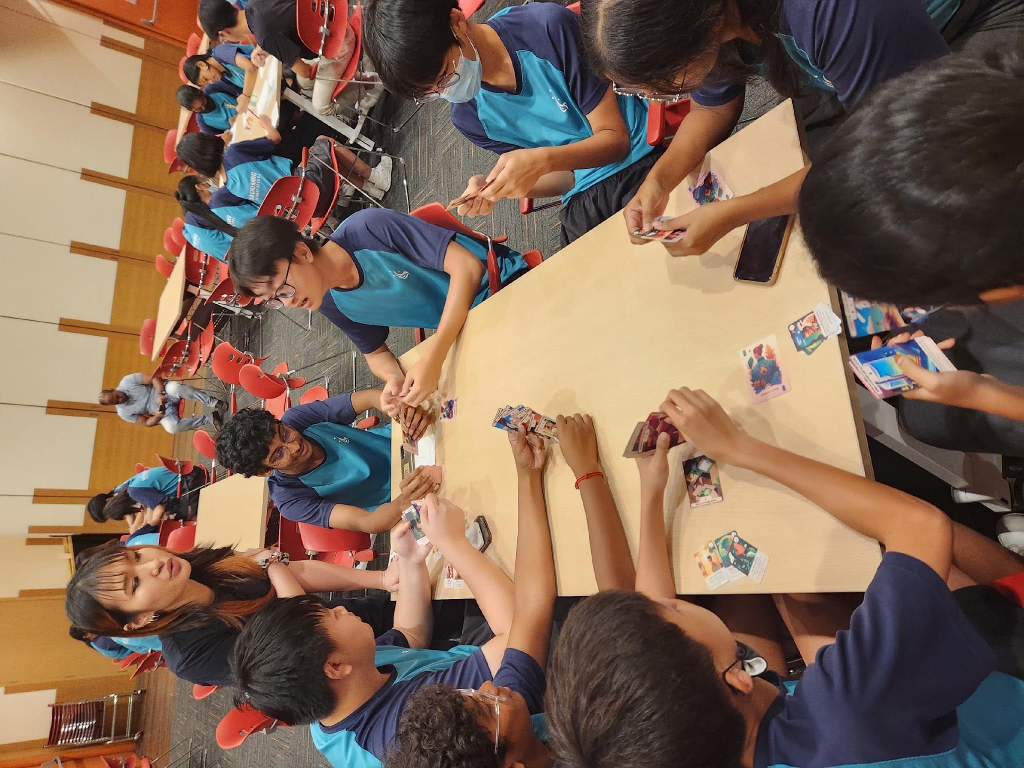 Students in blue uniforms gathered around a table playing a card game in a room with red chairs.