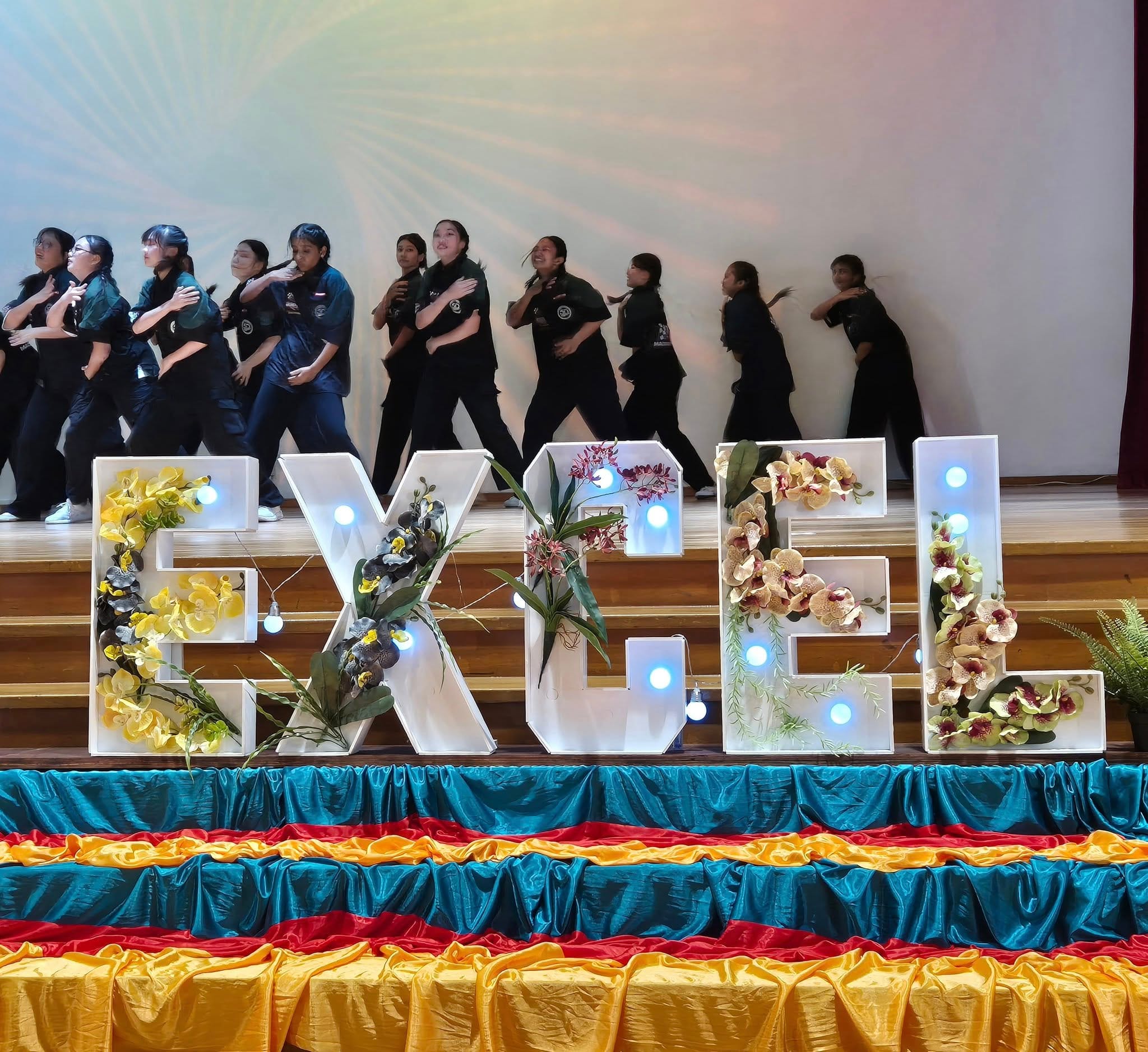 Students in black uniforms dance on stage behind lit letter signs spelling EXCEL decorated with flowers.