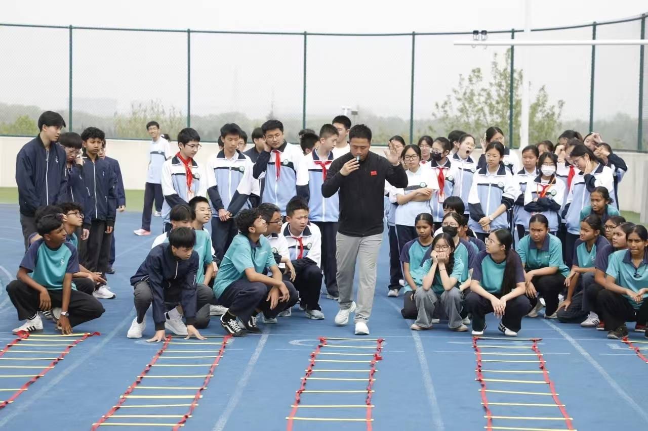 A coach addresses a large group of students on a blue outdoor track with agility ladders laid on the ground.