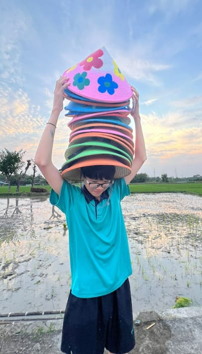Child in teal shirt balancing a tall stack of colorful cone-shaped hats on their head near a rice field.