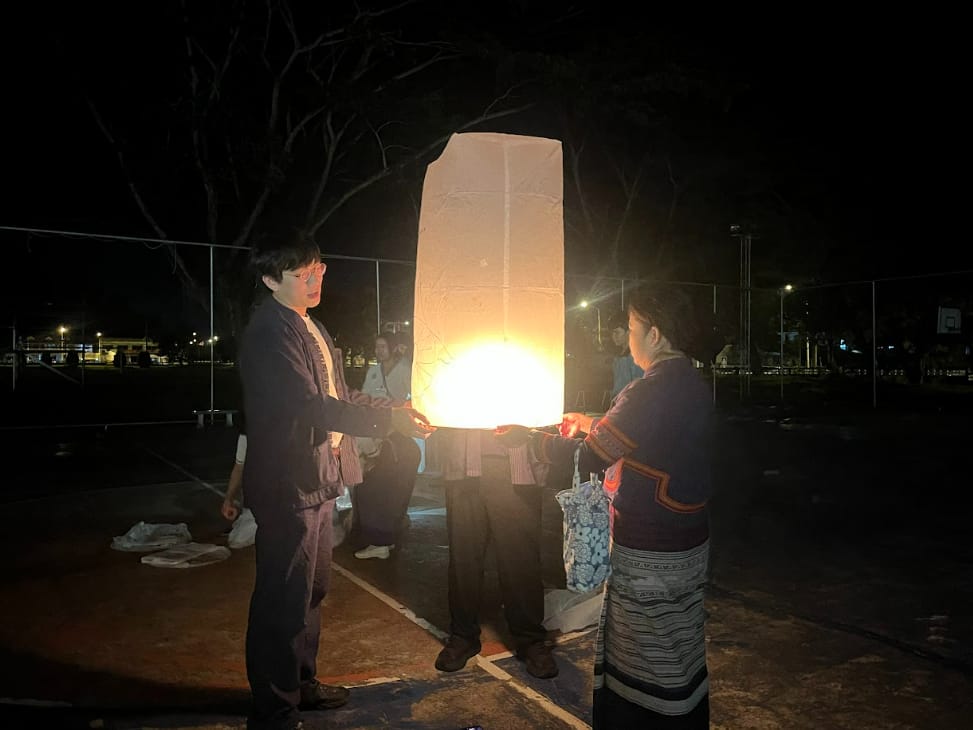 Three people in traditional clothing hold a glowing paper sky lantern at night in an outdoor park.