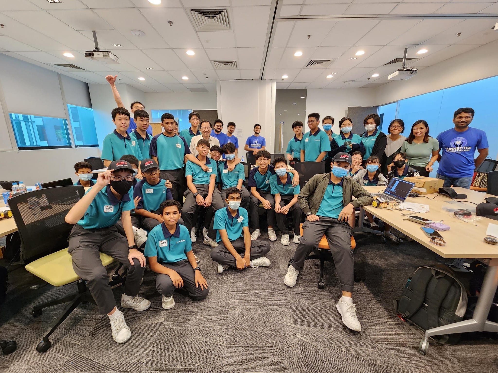 A large group of students in teal polo shirts and face masks pose together in a modern classroom setting.