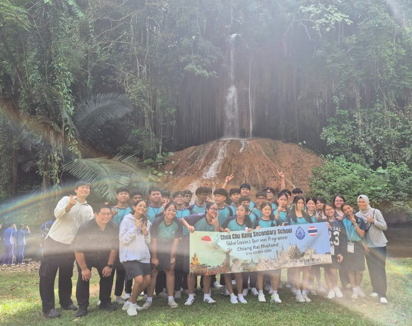 Group of students in teal shirts holding a Chua Chu Kang Secondary School banner in front of a waterfall.