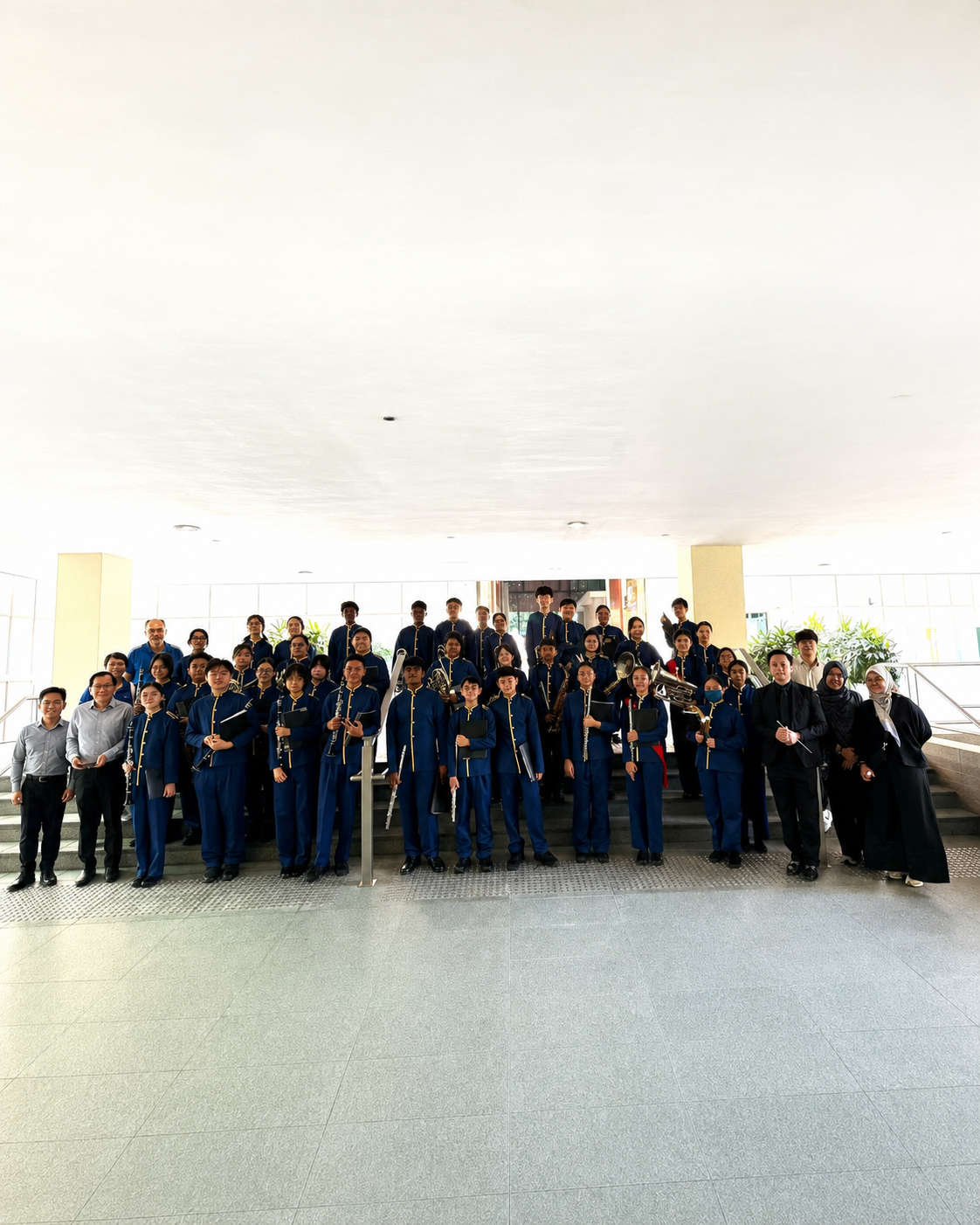 A group of students in blue stand with concert band instruments in front of a staircase