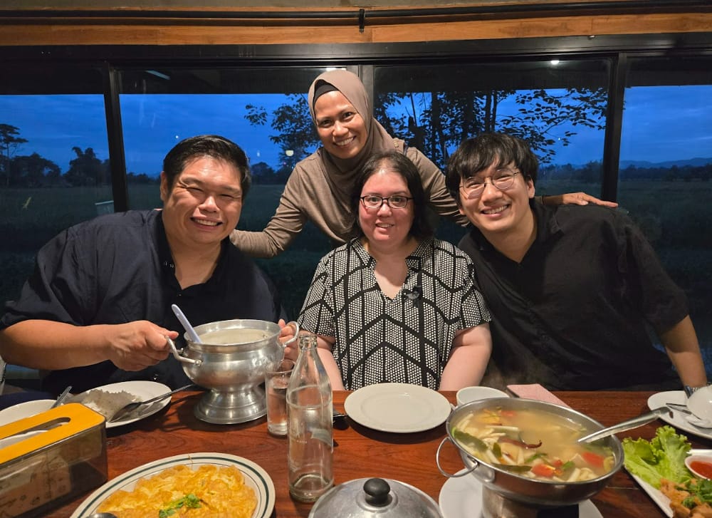 Four smiling friends pose at a dinner table with soup and food dishes, a dusk landscape visible through the window behind them.