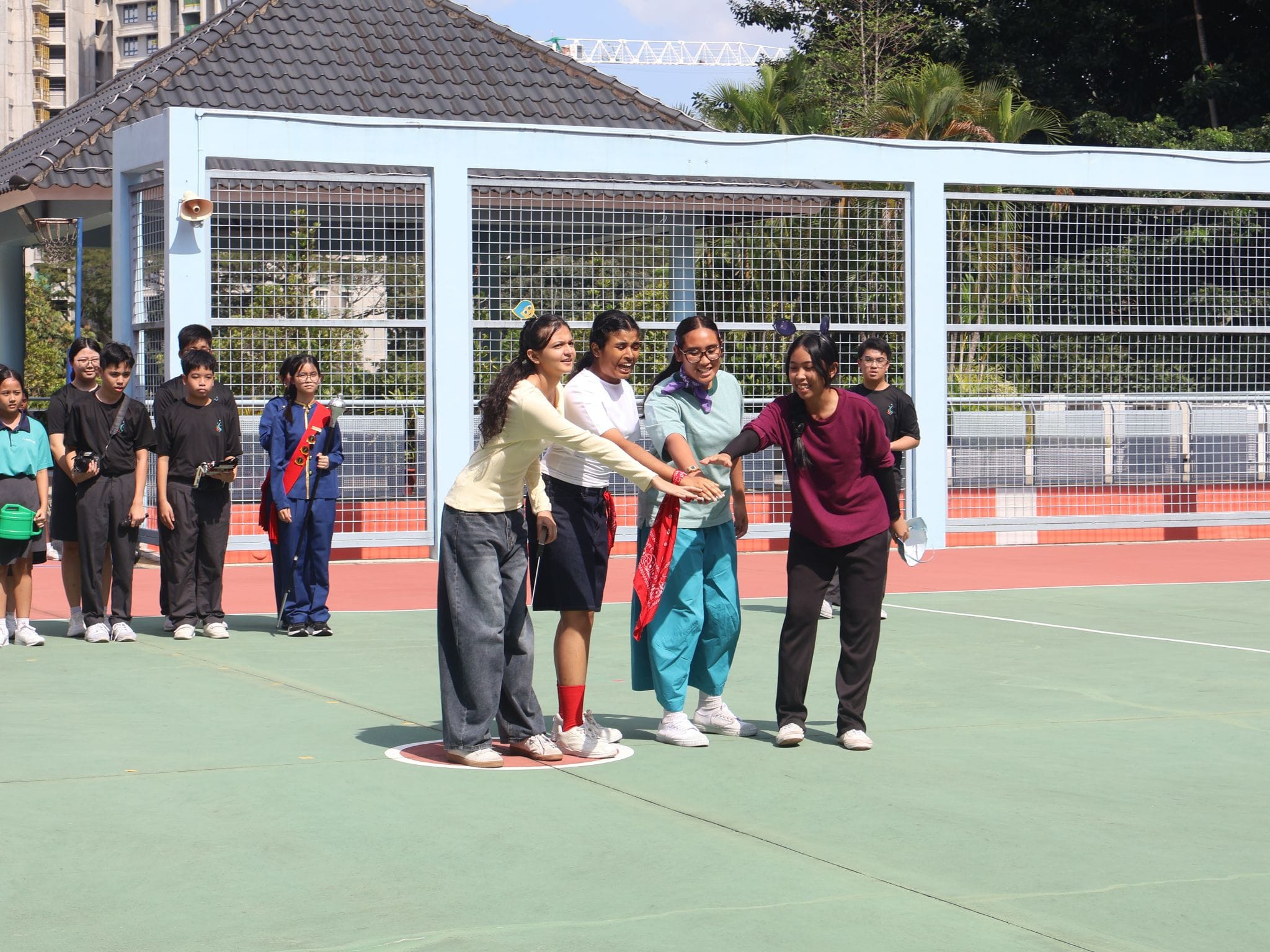 Four students join hands in a circle on an outdoor school court while classmates watch from the sideline.