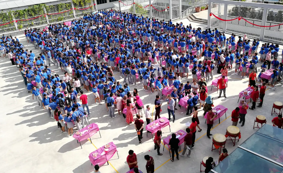 Large crowd of students in blue uniforms gathered in an outdoor courtyard with pink tables and drums nearby.
