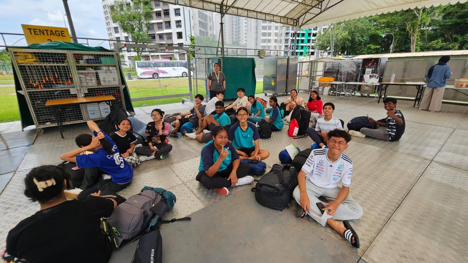 A group of teenagers sitting on the ground under a large outdoor tentage with backpacks, posing for the camera.