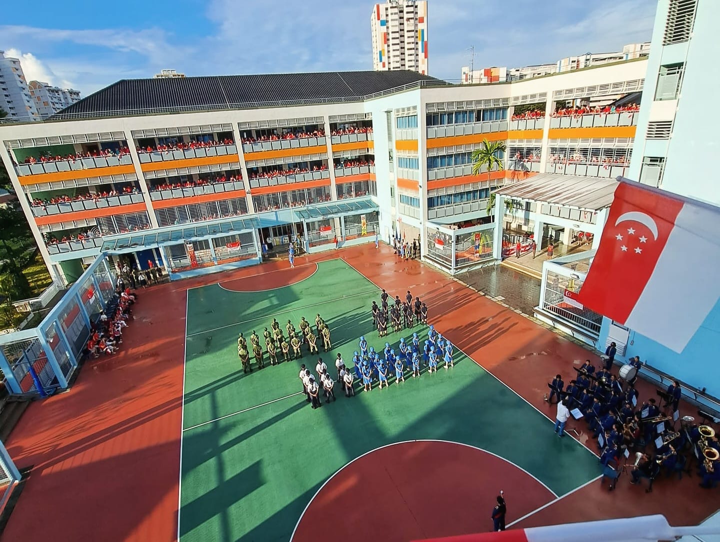 School courtyard ceremony with uniformed students on a basketball court, a Singapore flag hanging, and spectators on balconies.