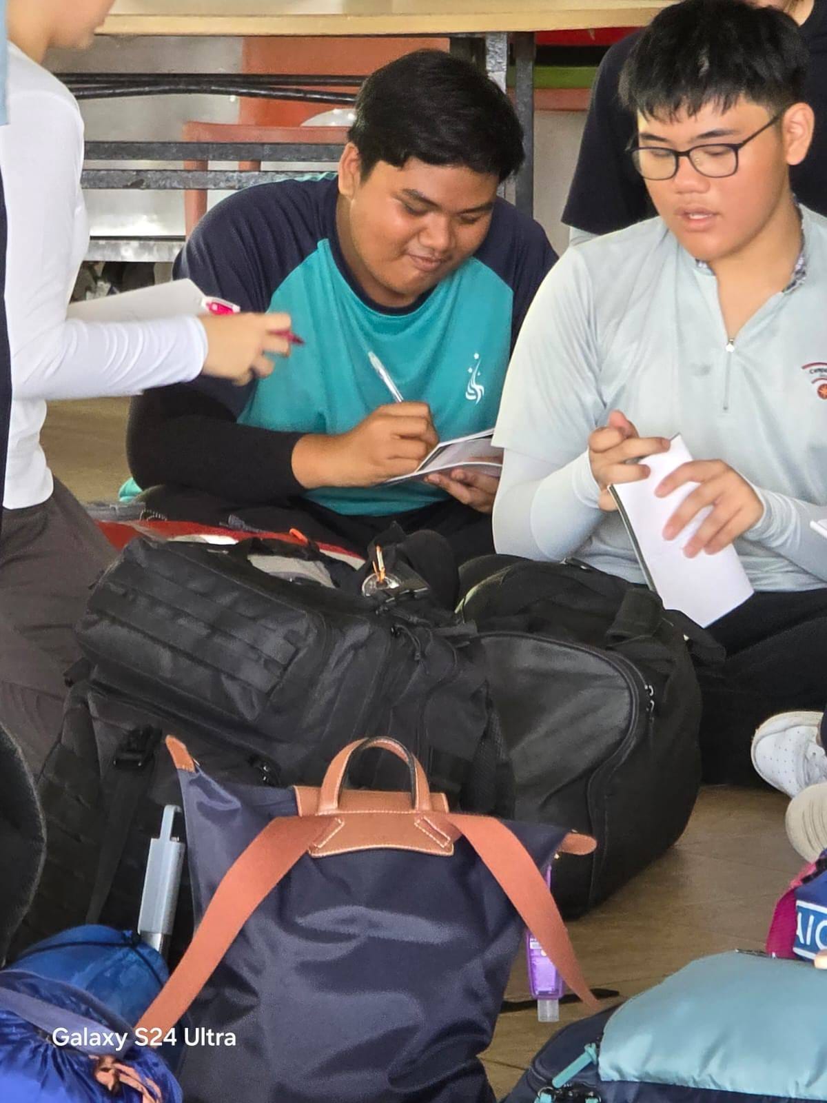 Two teenage boys sit on the floor with bags, one writing in a booklet and the other holding papers.