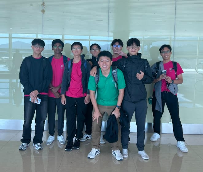 Eight young men pose together indoors, most wearing pink shirts, one in green, standing on a tiled floor.