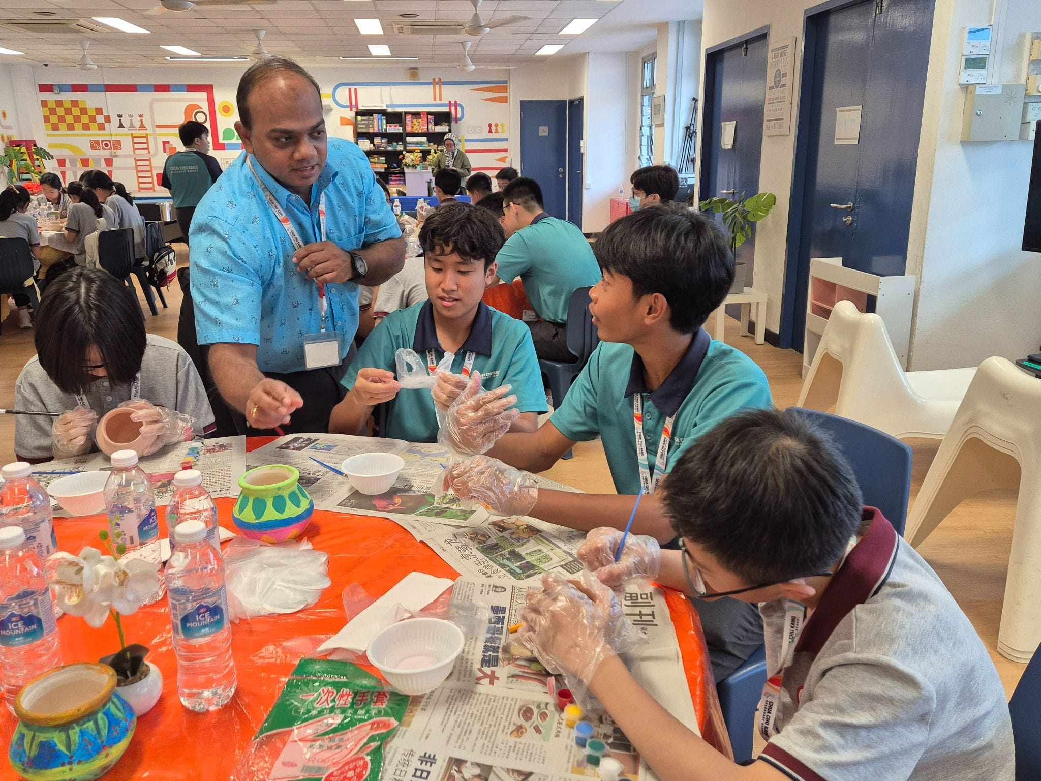 A teacher in a blue shirt supervises students wearing gloves painting clay pots at a table covered with newspaper.