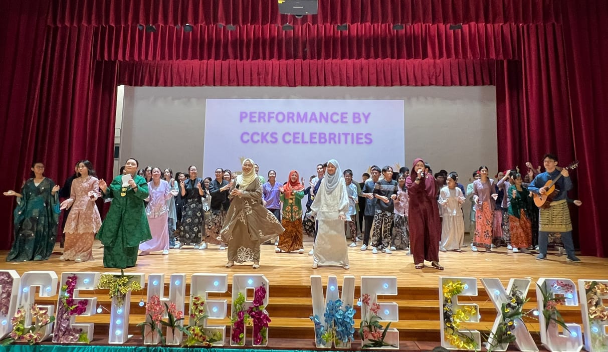 A large group in colorful traditional outfits performs on a stage with a screen reading "Performance by CCKS Celebrities."