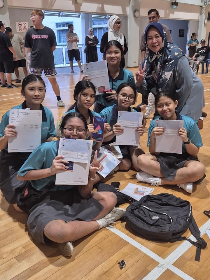 Six female students in teal uniforms sit on a gym floor holding papers, posing with a smiling adult woman behind them.