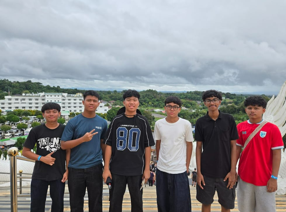 Six teenage boys posing together on outdoor steps with a town, green trees, and cloudy sky in the background.