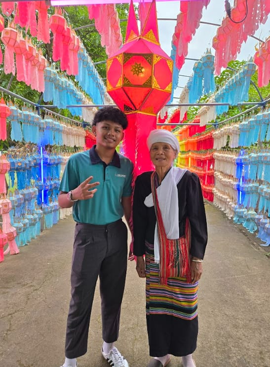 A young man and an elderly woman pose under rows of colorful hanging lanterns at a festive outdoor display.