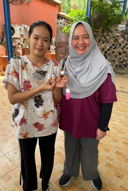 Two women smiling outdoors, holding small crocheted figurines, one wearing a gray hijab and purple top.