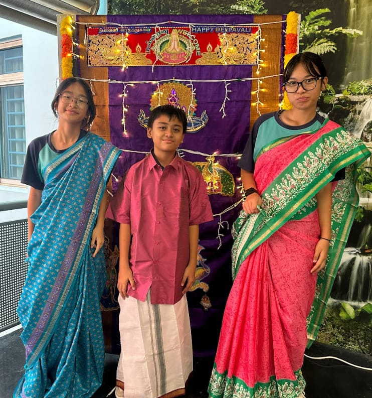 Three children in traditional Indian attire pose before a decorated Happy Deepavali banner with fairy lights.