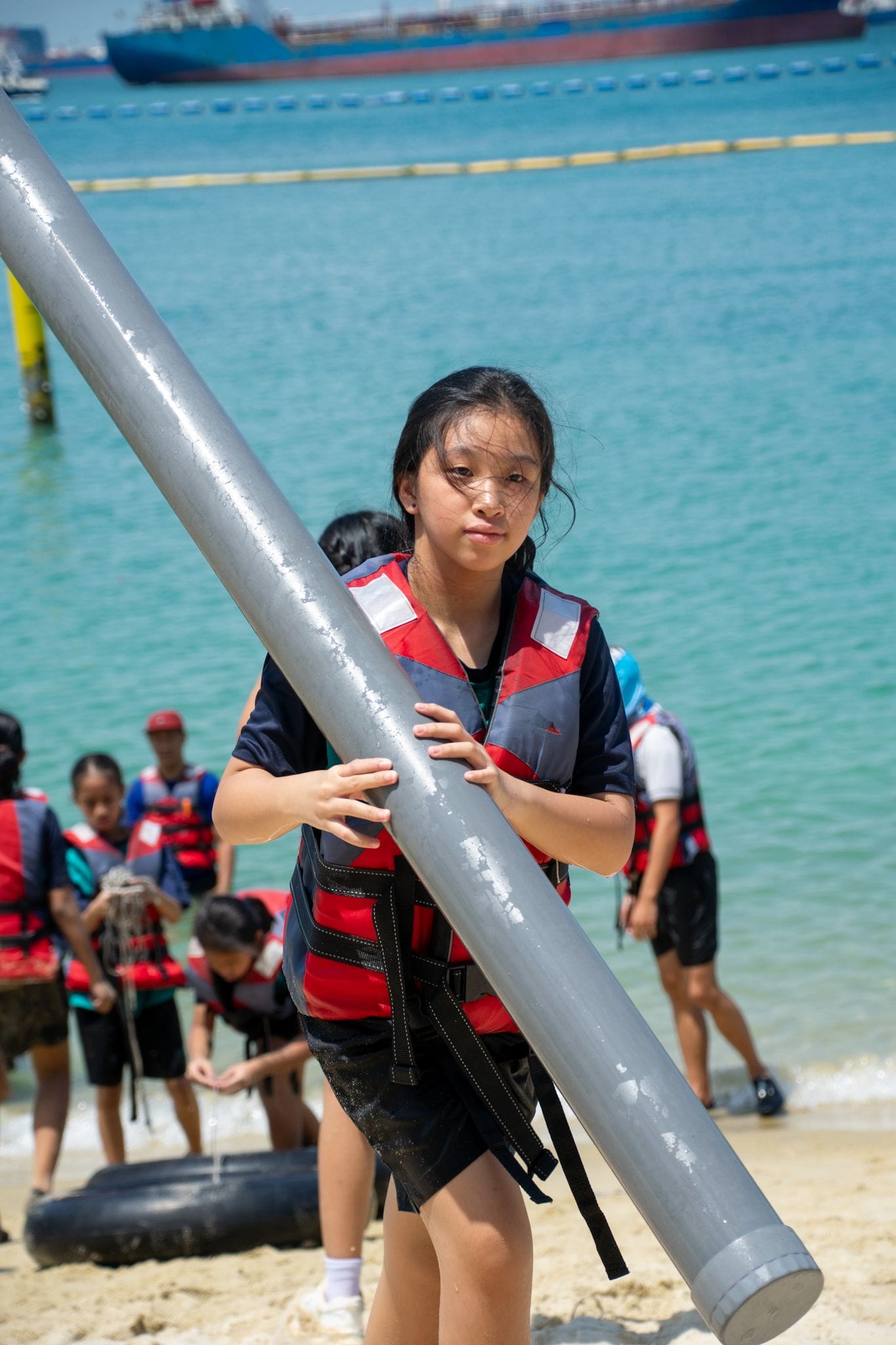 A girl in a red life vest holds a large metal pole on a sandy beach with other children and ships in the background.