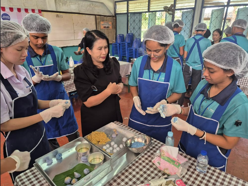 A group wearing blue aprons and hair nets shapes dough balls at a table during a food preparation class.