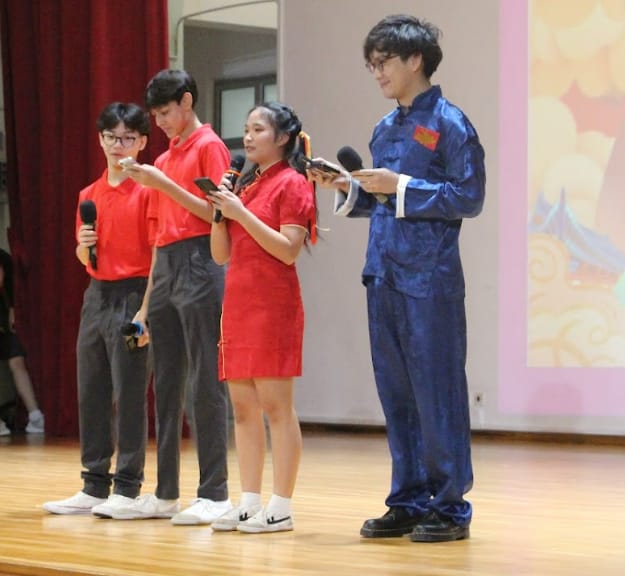 Four students on a stage holding microphones, three in red outfits and one in a blue traditional suit.
