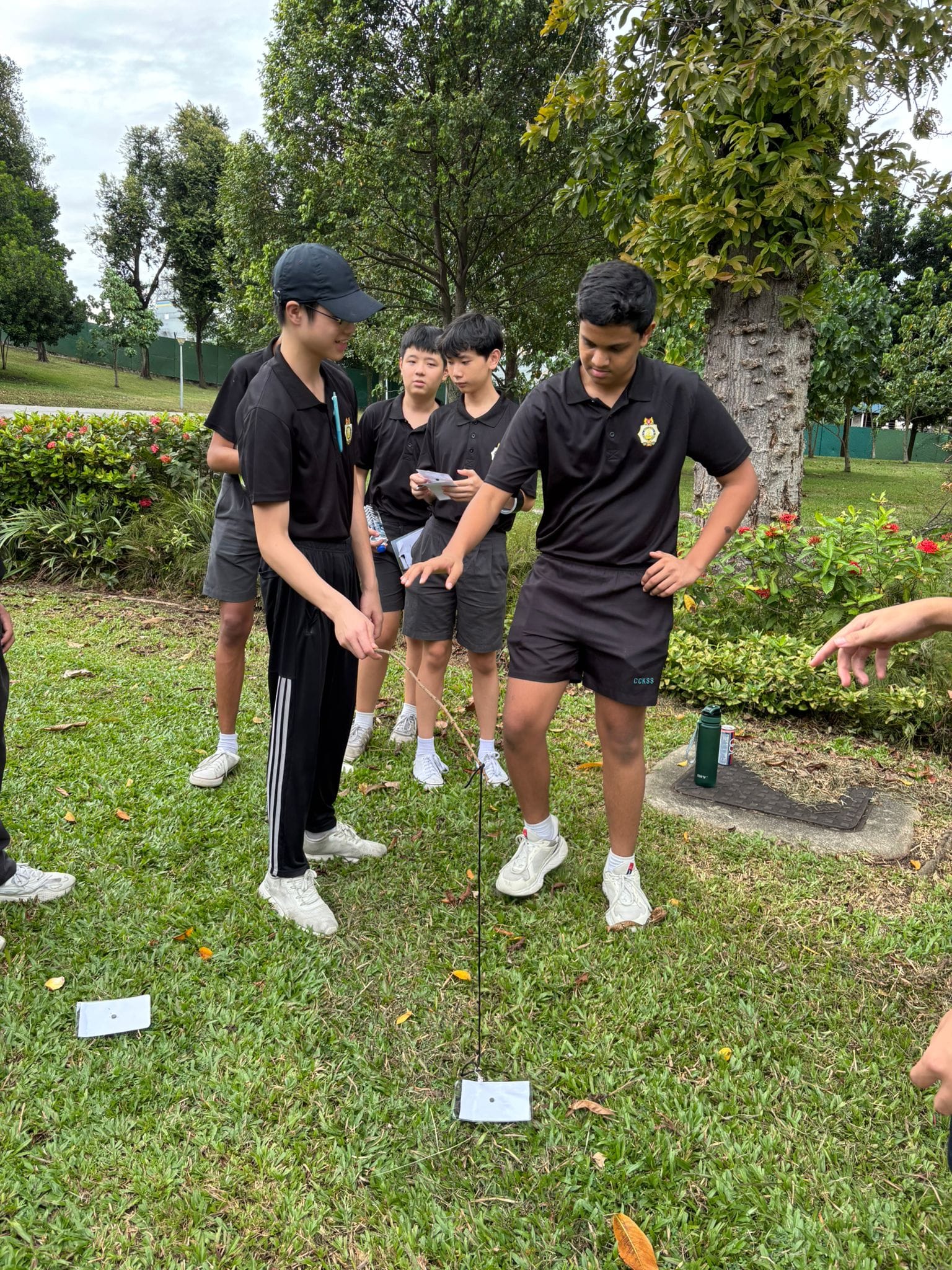 Four students in black uniforms stand on grass outdoors, examining a string or wire anchored to the ground.