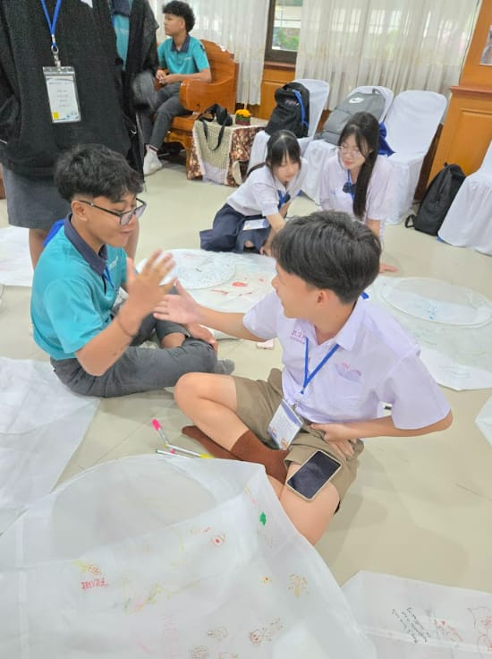 Students in uniforms sit on a floor surrounded by large paper sheets with drawings, engaged in discussion.