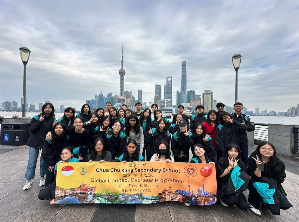 A group of students in black and teal jackets pose with a Chua Chu Kang Secondary School banner in front of the Shanghai skyline.