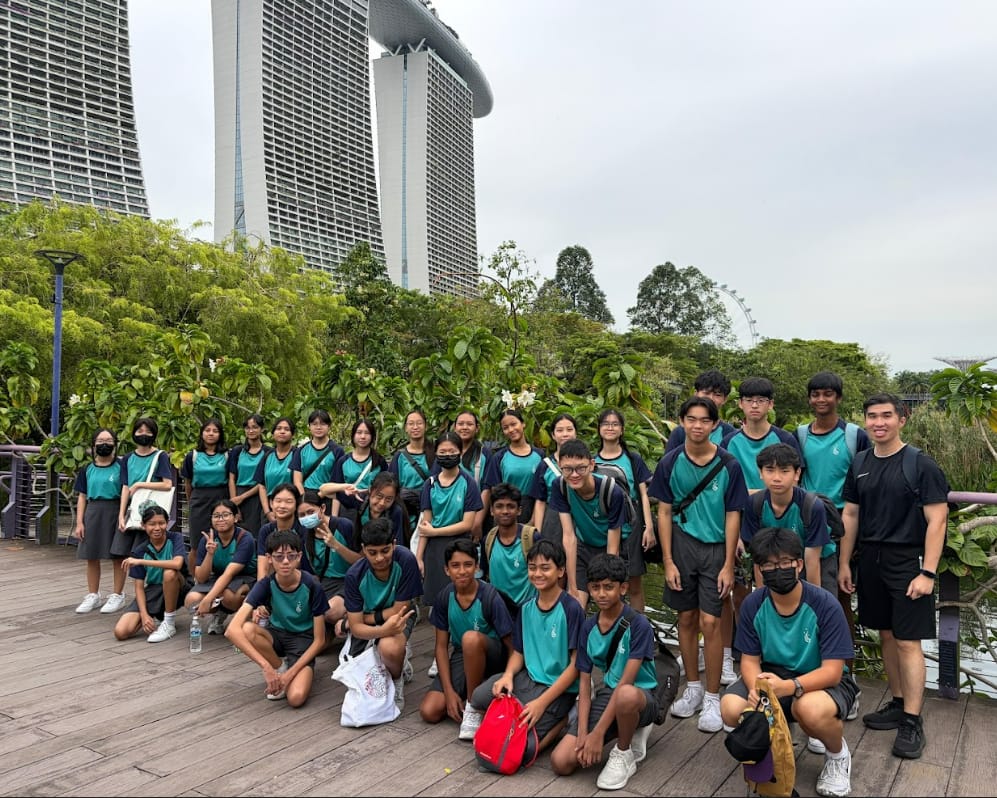 A large group of students in teal uniforms pose on a wooden walkway with Marina Bay Sands towers behind them.