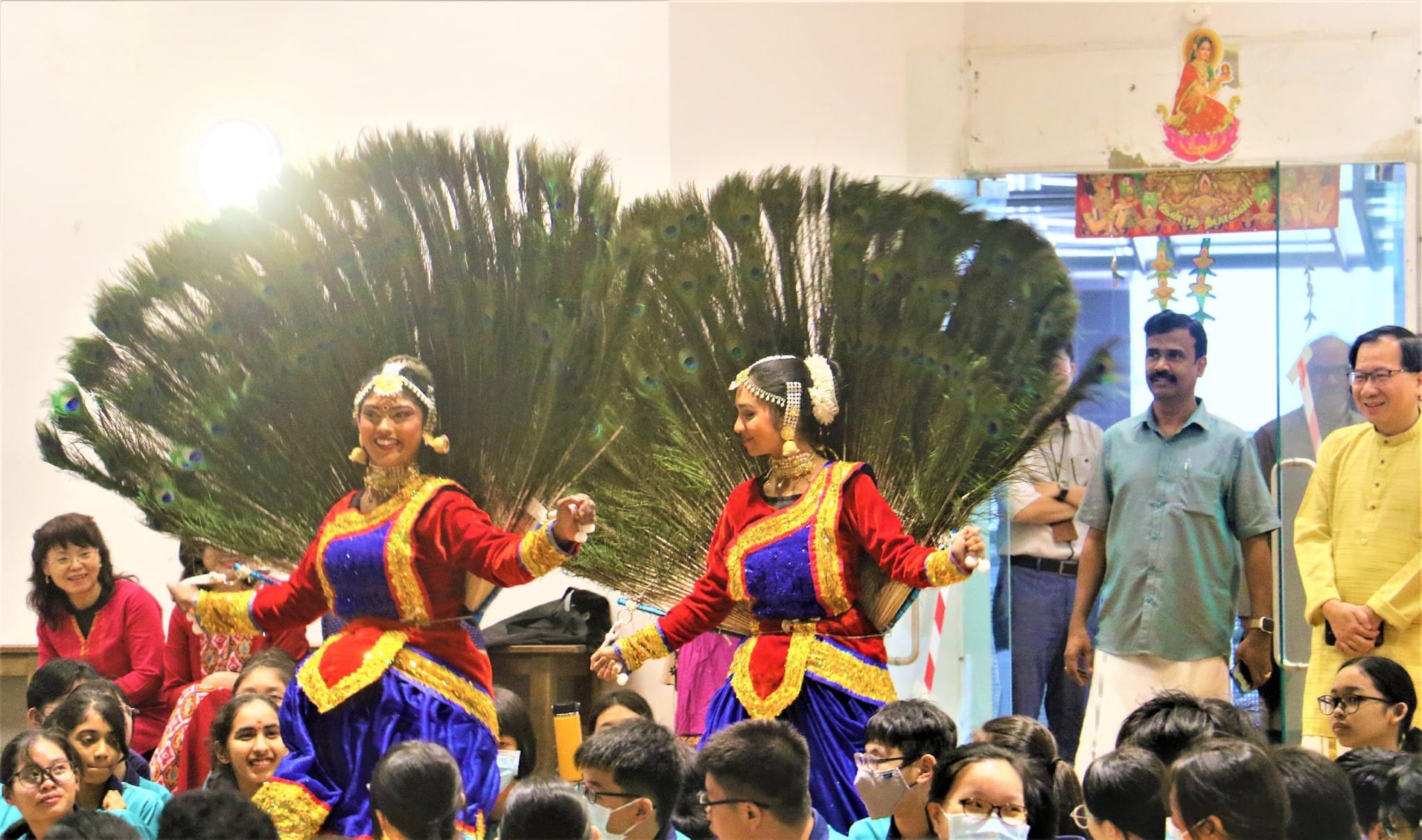 Two women in red and blue Indian dance costumes perform with large peacock feather fans before a seated crowd.