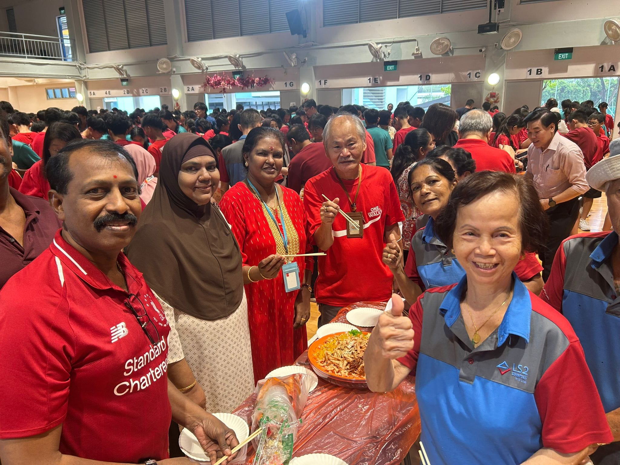 A diverse group of smiling adults gathers around a table with food at a crowded indoor community event.
