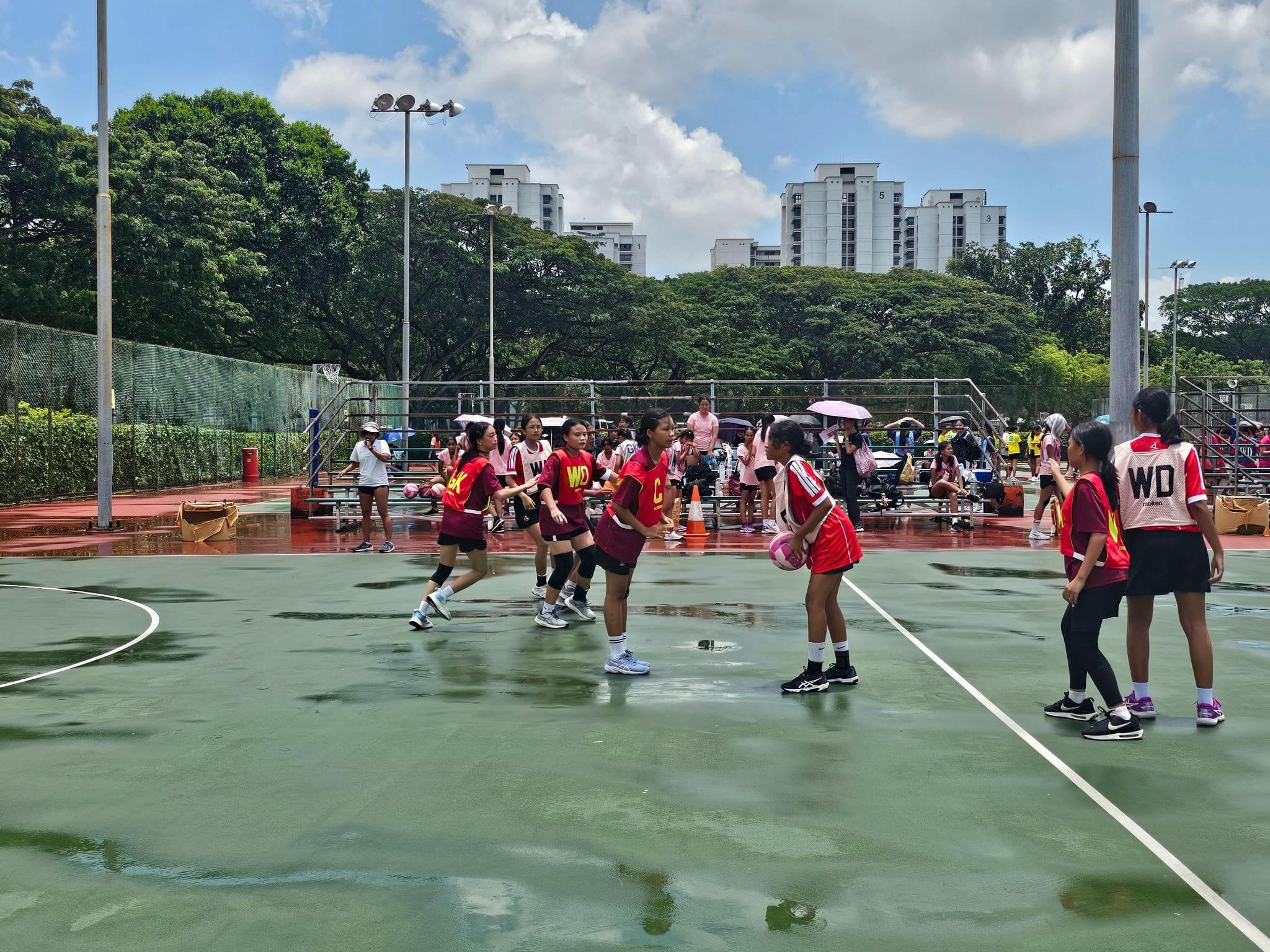 Girls in red uniforms play netball on a wet outdoor court with spectators, trees, and apartment buildings in the background.
