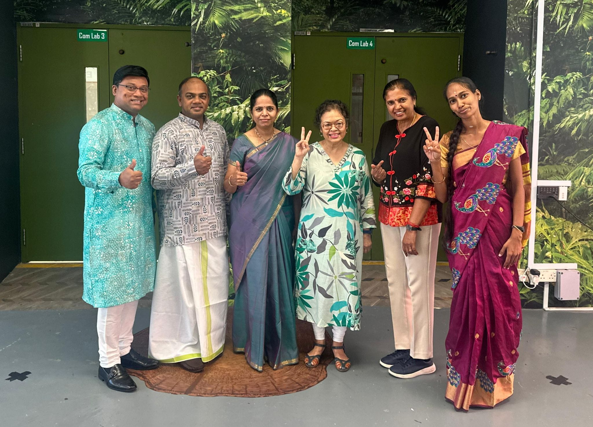 Six people in traditional Indian attire smile and pose with thumbs up and peace signs outside computer labs.