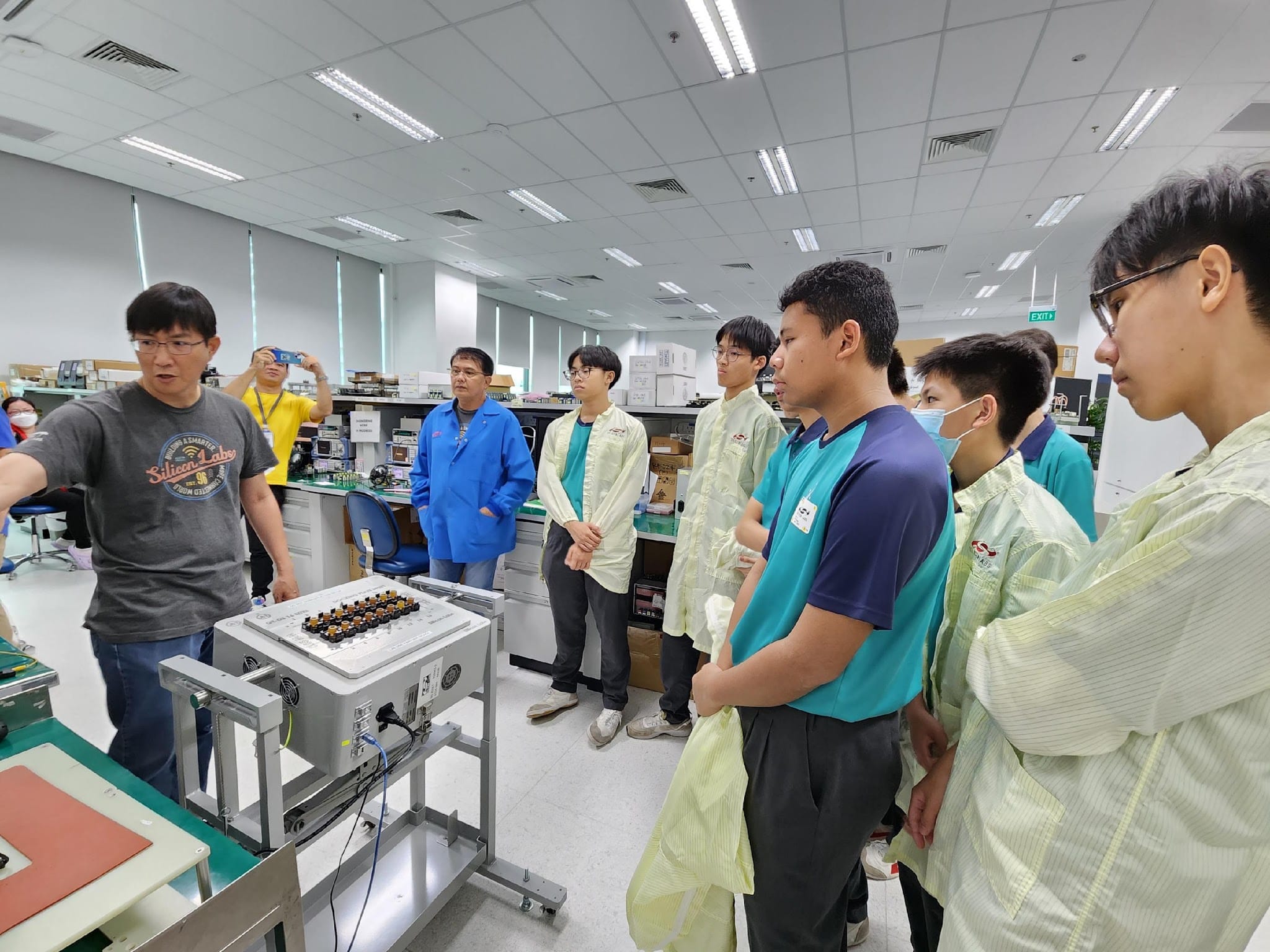 A man presents electronic equipment to a group of students in lab coats inside a bright technical facility.