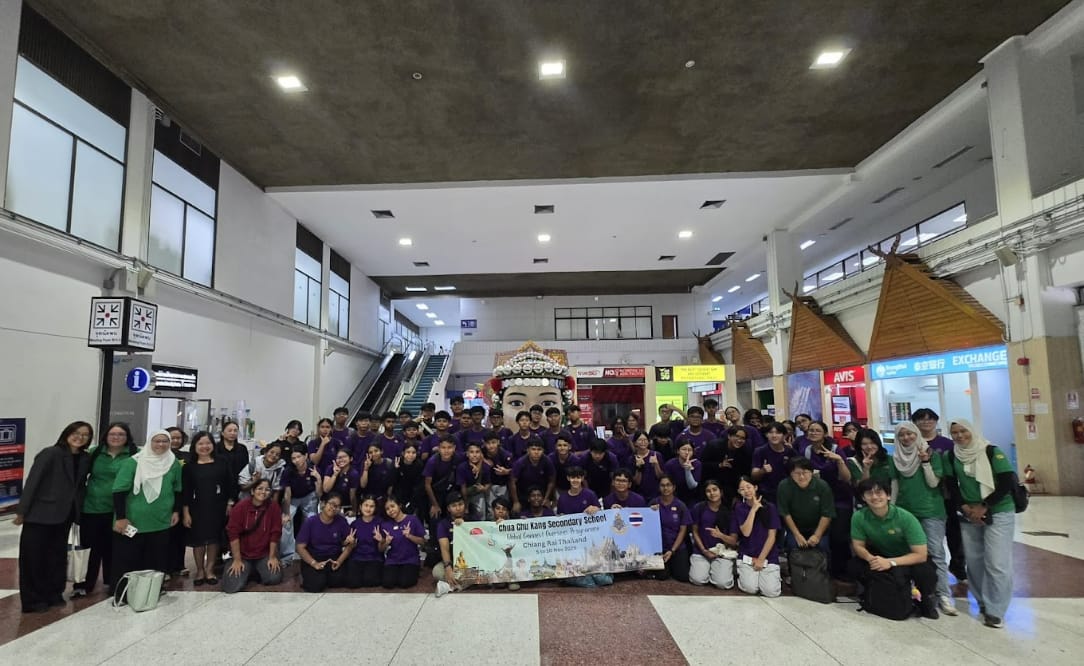 Large student group in purple and green shirts poses with a banner inside Chiang Rai Thailand airport.