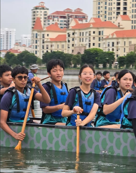 A group of young people wearing blue life jackets paddle a green dragon boat on a river with buildings behind.