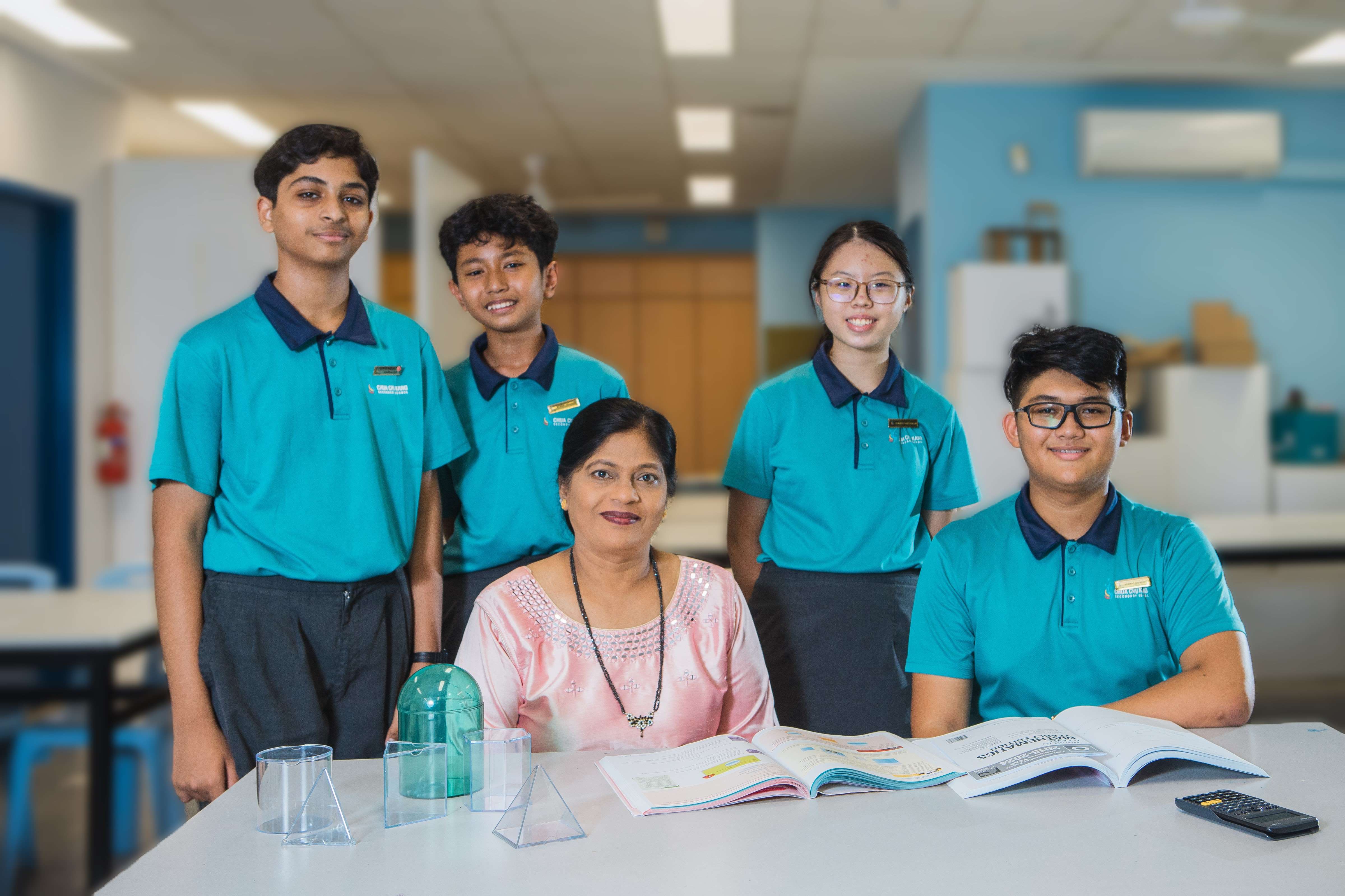 A teacher in a pink top sits at a desk with four students in teal uniforms in a school classroom.