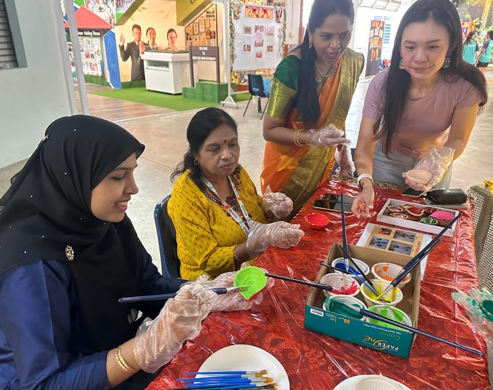 Four women in traditional attire do a painting craft activity at a red table with colorful paints and brushes.