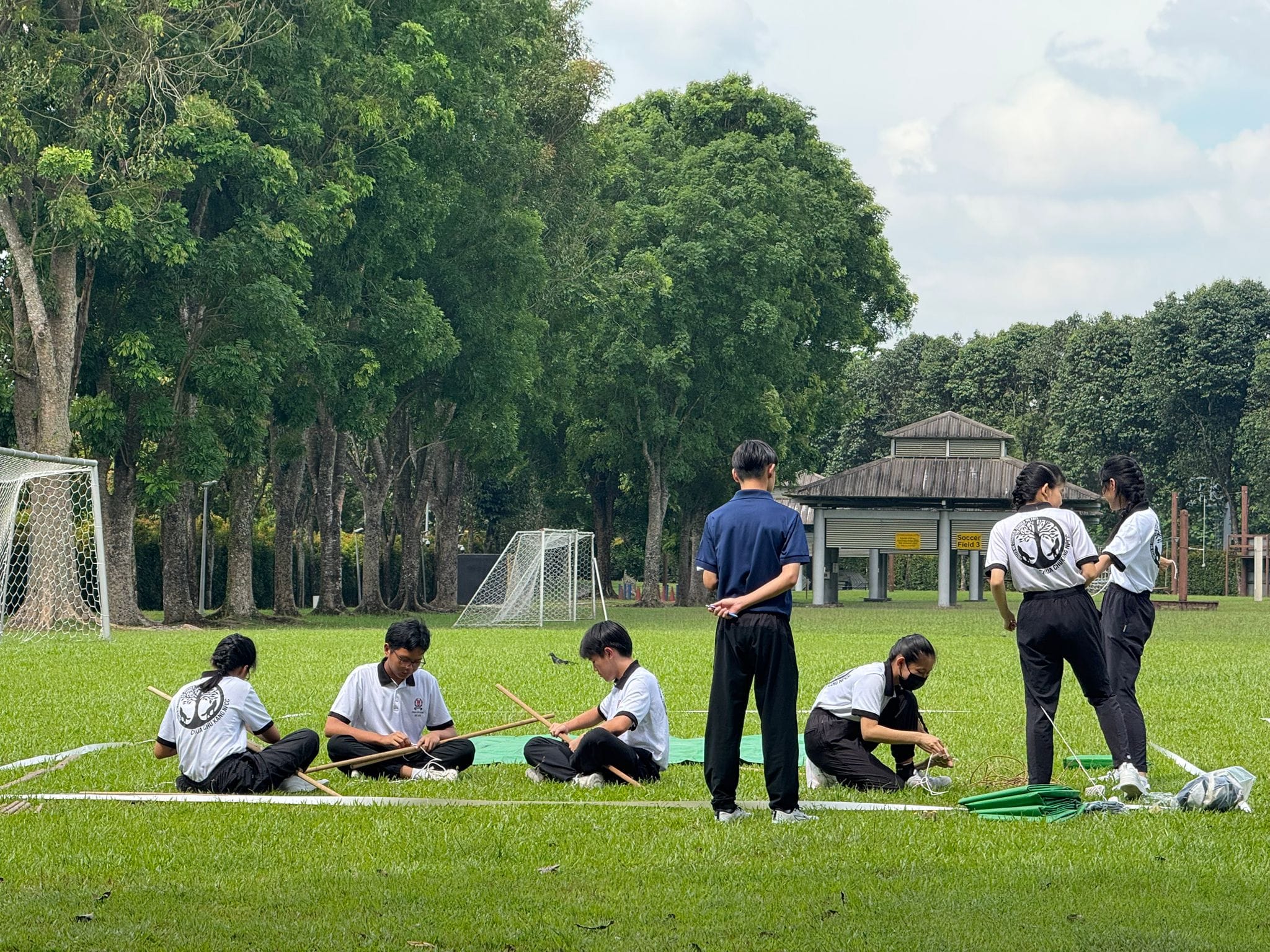 Students in uniforms sit and stand on a grass field assembling wooden poles and ropes near a soccer goal.