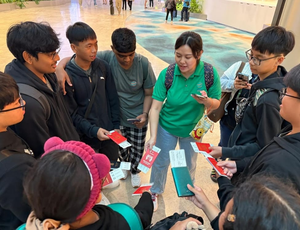 A woman in a green shirt stands in a circle with several young people, all holding red booklets and tickets indoors.