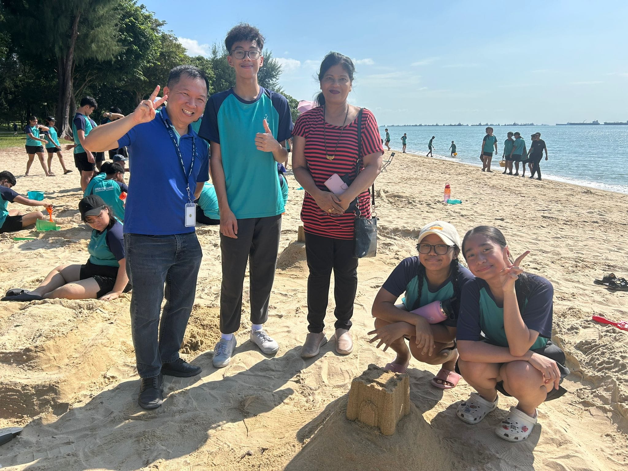 Five people pose on a sunny beach near a sand sculpture, with a group of students in teal shirts visible behind them.