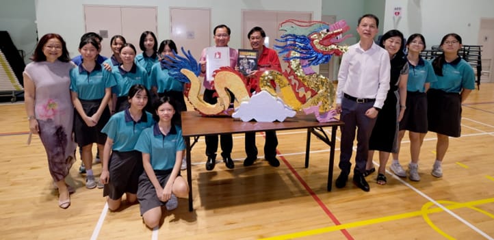 A group of students and adults in teal uniforms pose around a colorful dragon sculpture on a table in a gym.